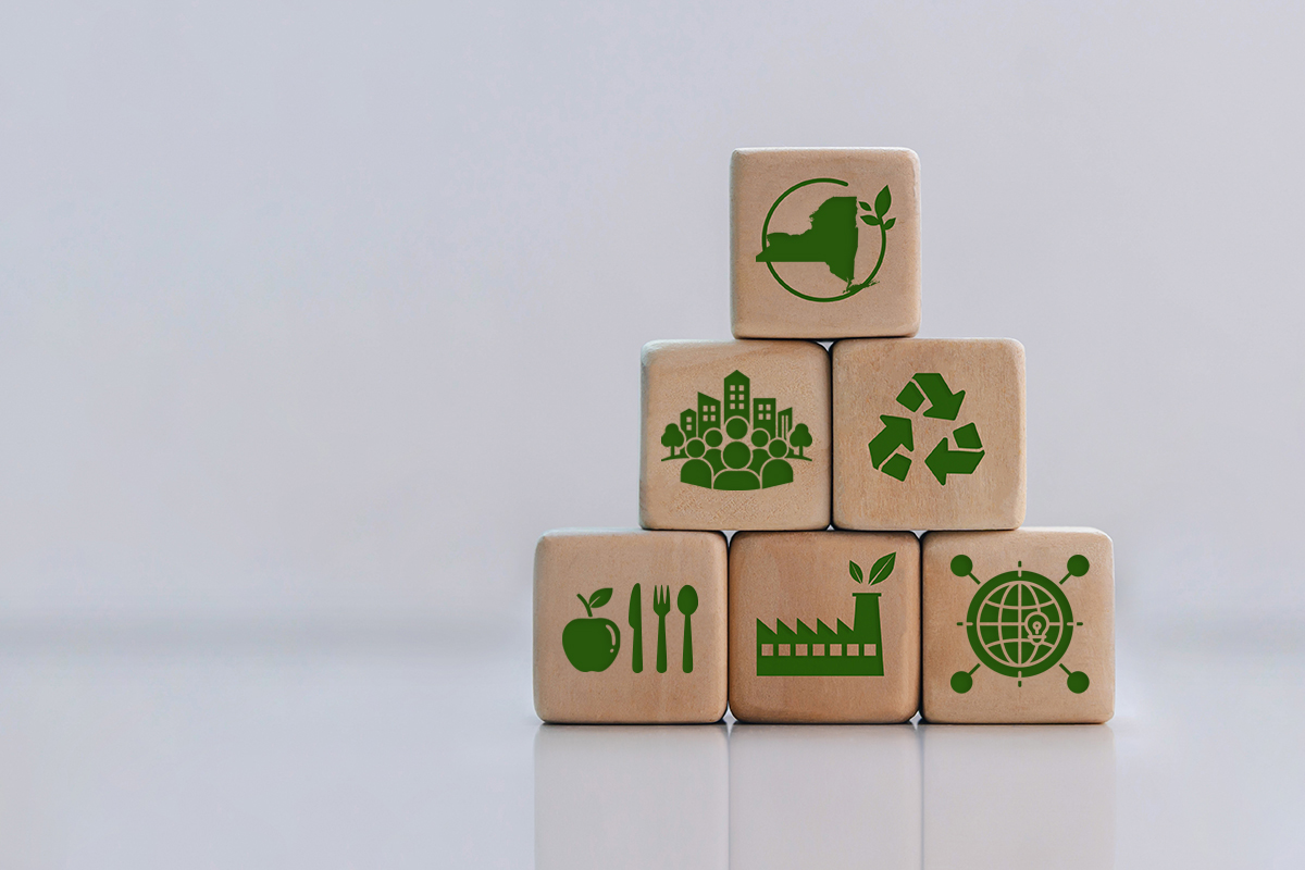 Six wooden blocks stacked in a pyramid, printed with green icons for New York State, community, recycling, food, clean manufacturing, and global innovation.